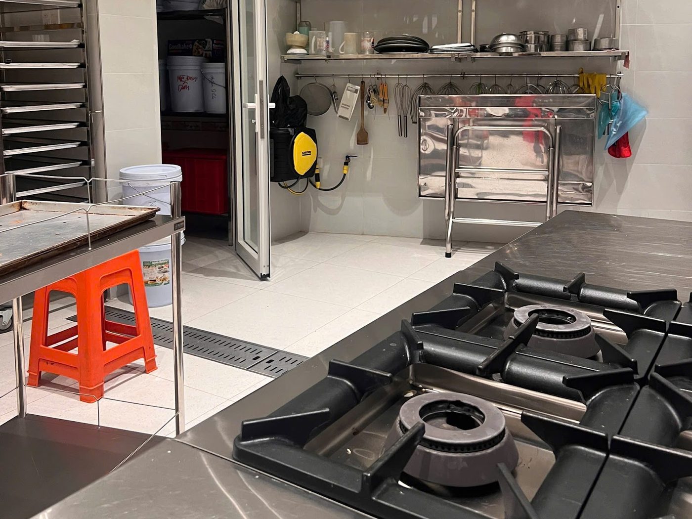Kệ inox treo tường cùng thanh móc dụng cụ đặt ngay trên tuyến thao tác của xưởng bánh, bếp gas công nghiệp phía trước