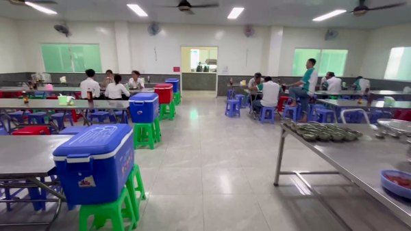 Phòng ăn bếp ăn công nghiệp: bàn dài ghế nhựa, thùng nước kê trên ghế, khay chờ đặt ở lối đi