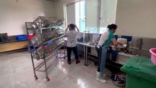 Khu rửa bếp ăn công nghiệp: nhân viên rửa thủ công tại chậu, xe kệ khay đứng cạnh thùng rác